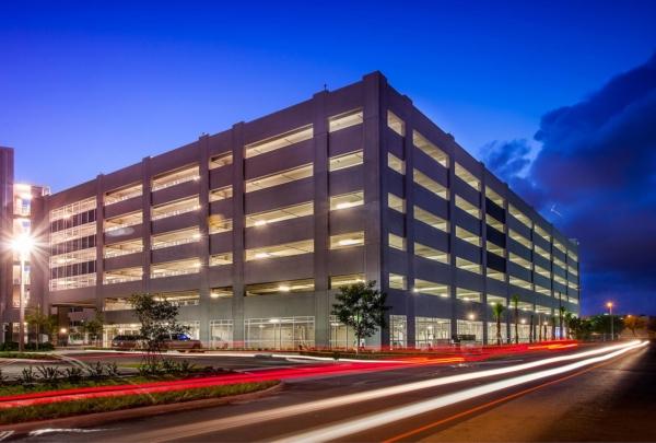 Exterior photo of the Miami Dade College Hialeah Campus parking garage. Evening photo with bright blue skies. Garage lights on. Vehicle lights create streaks on the adjacent street.